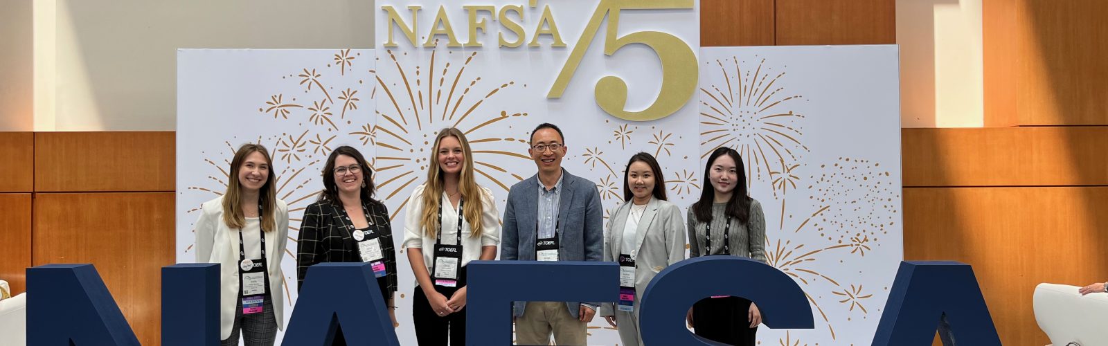 A group of 6 individuals stands by large NAFSA letters at a professional conference.